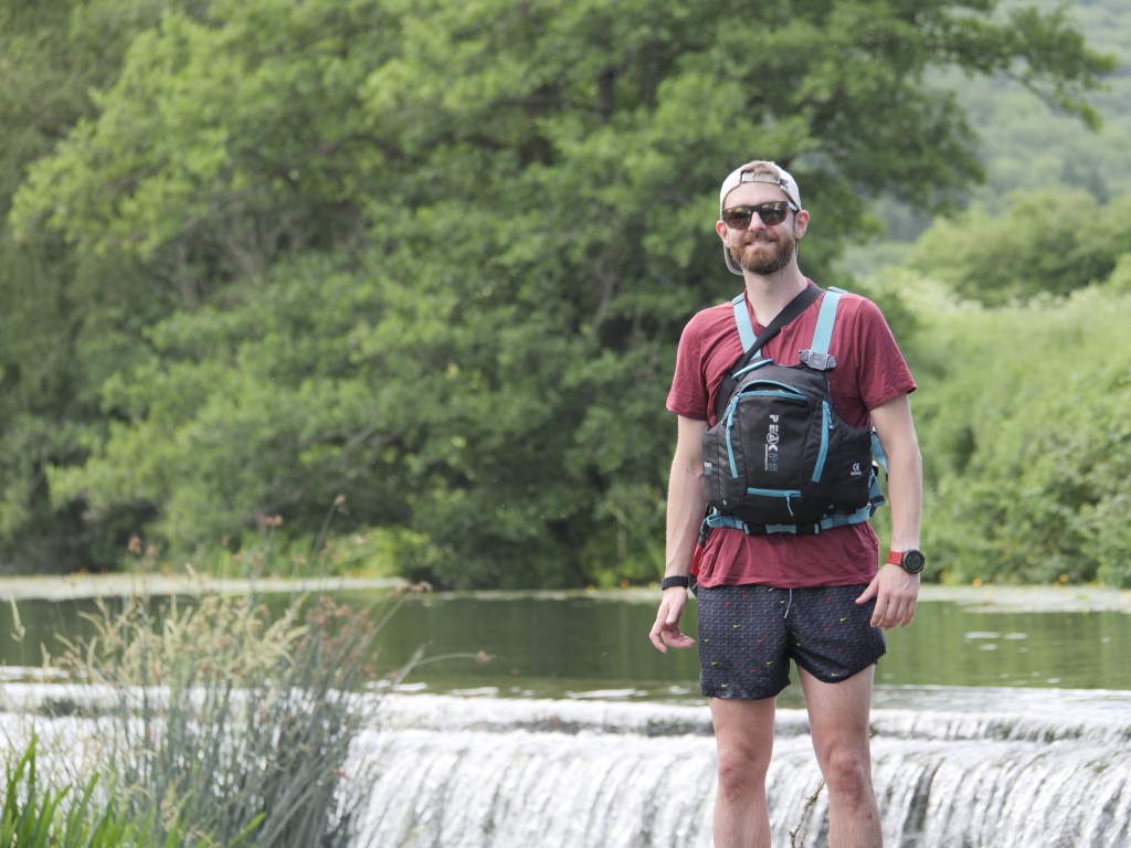 A man wearing sunglasses and a red t-shirt stands by a river with a small waterfall in the background, dressed in shorts and a backpack, surrounded by greenery.