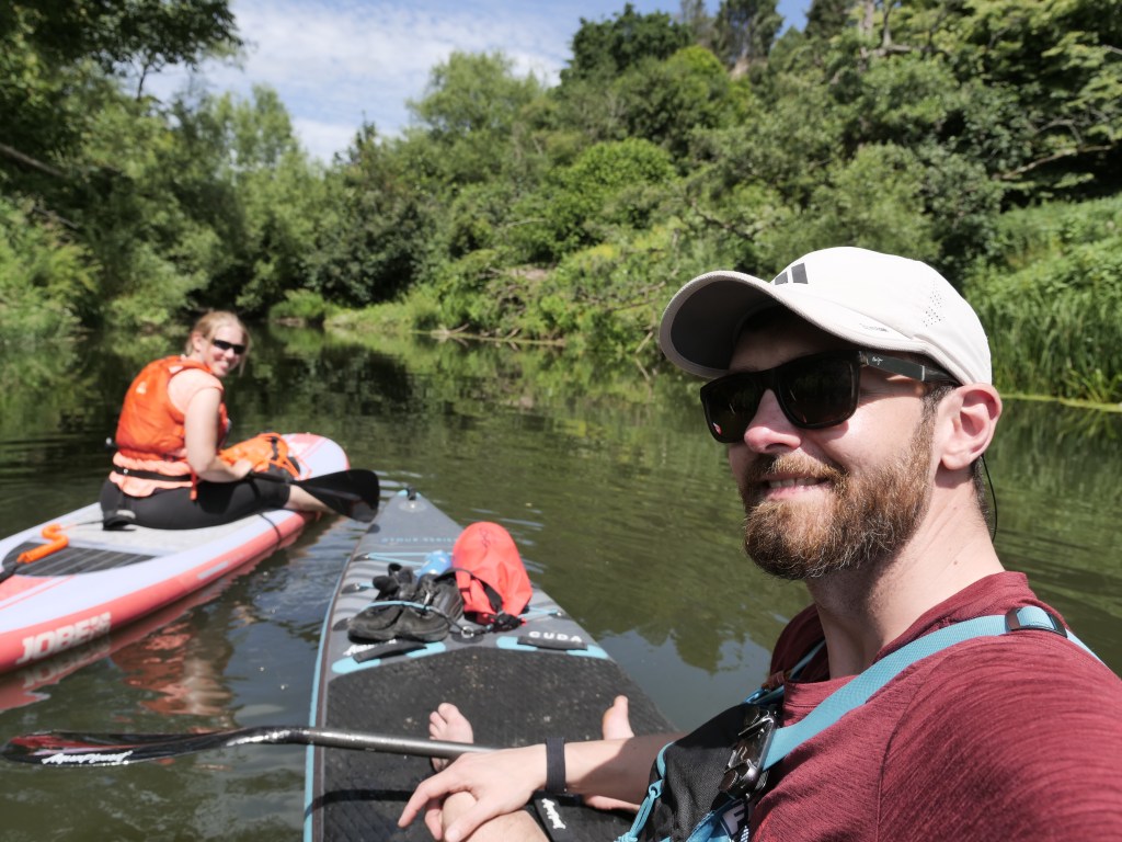 Two adults enjoying a kayaking trip on a calm river, with lush greenery in the background. One person is on a stand-up paddleboard, while the other takes a selfie in a kayak.