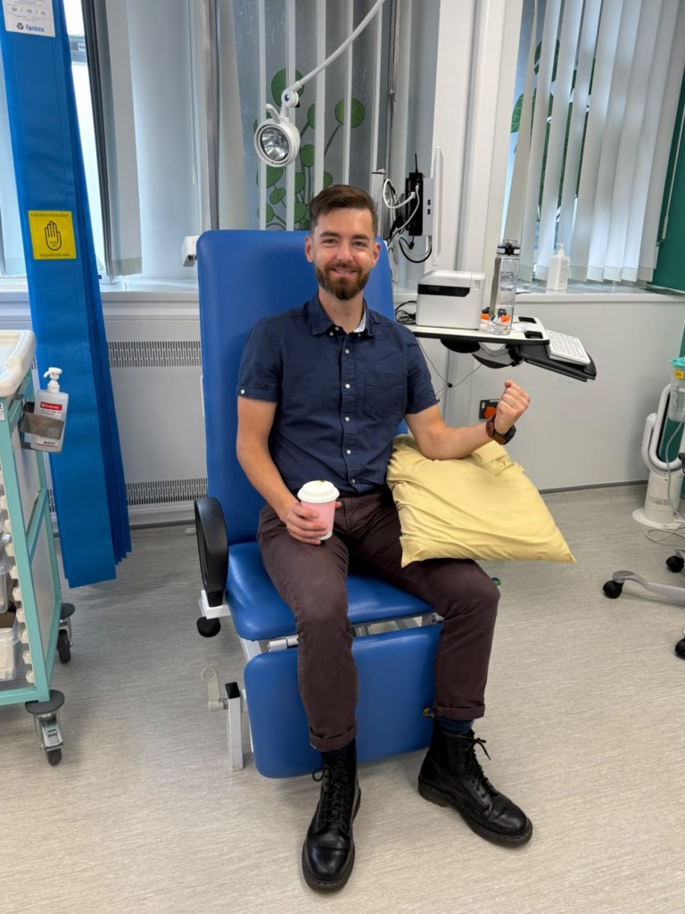 A man sitting in a clinical setting on a blue chair, holding a coffee cup and a cushion. He is wearing a dark shirt and black boots, smiling towards the camera.