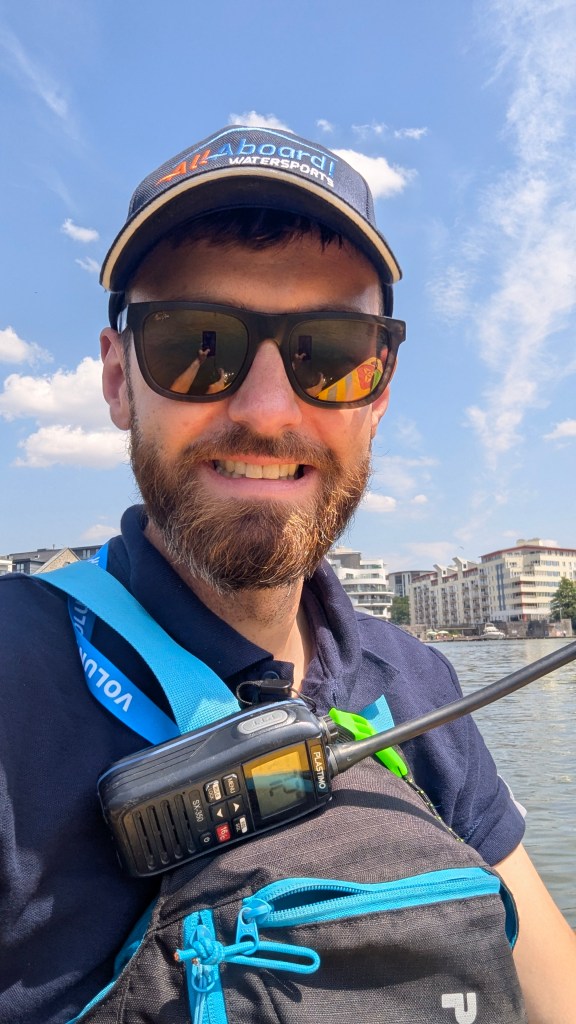 A man wearing sunglasses and a cap smiles at the camera while holding a radio on his chest, with a river and buildings in the background under a blue sky.