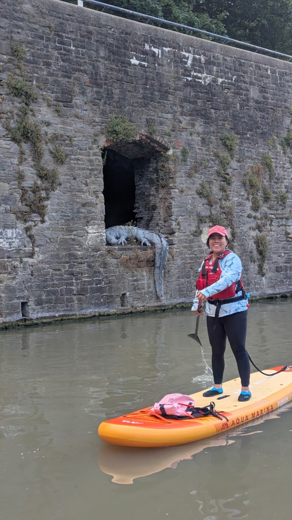 A person standing on a paddleboard in a canal, wearing a life jacket and a cap, with a stone wall in the background featuring a partially obscured opening.