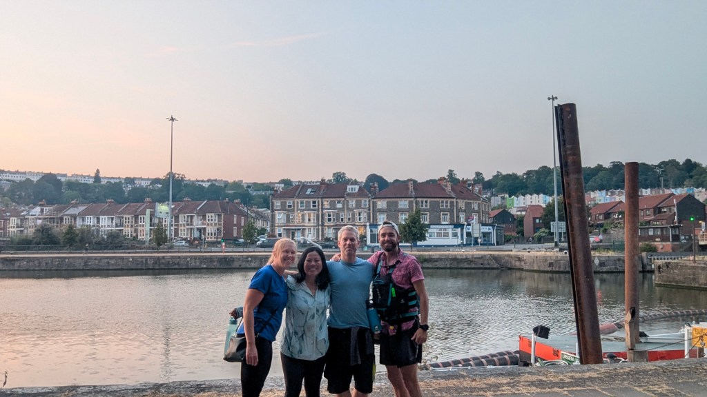 Four friends posing together by a river at sunset, with houses in the background and boats on the water.