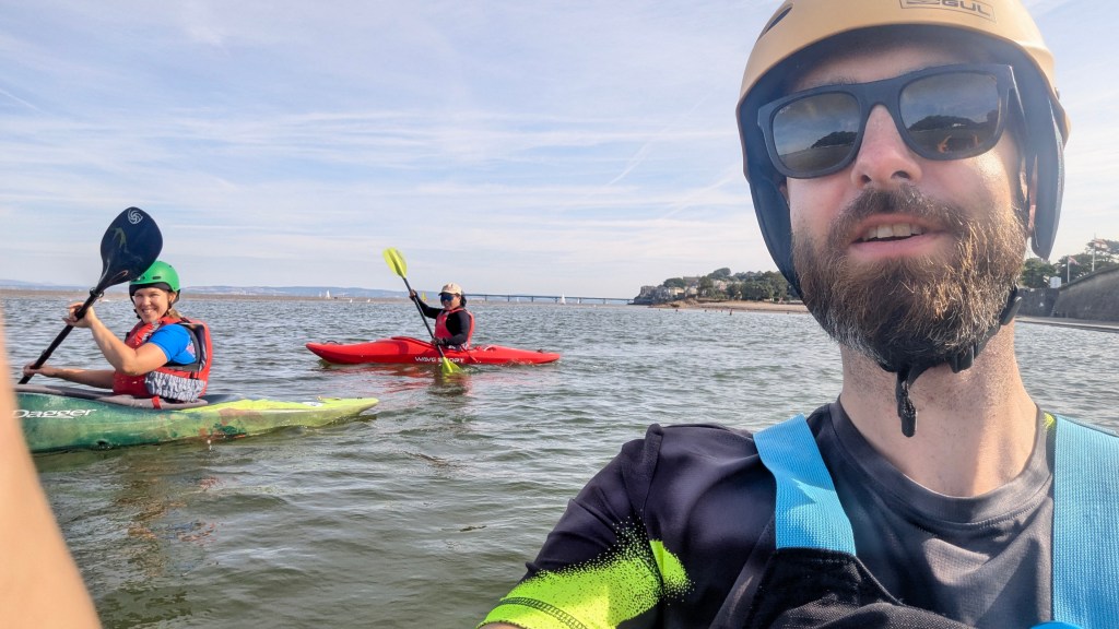 Three people kayaking on a calm water body, with one person in the foreground taking a selfie. The person on the left is in a green kayak, wearing a helmet and smiling, while the person in the red kayak on the right is paddling. The background features a sandy shore and a distant bridge.